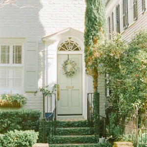 Charming front door with a wreath, surrounded by greenery and a white brick facade.