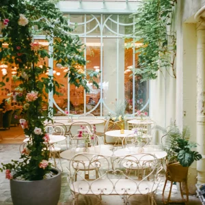 light-filled space with white wrought-iron chairs and tables, surrounded by potted plants and pink flowers under a glass ceiling.