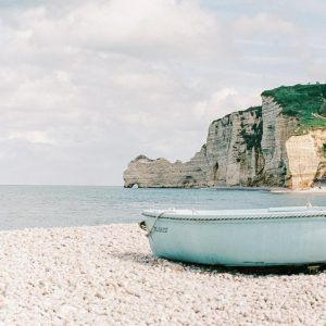 Pebble beach with a pale blue boat in the foreground, facing the calm sea and dramatic white cliffs of Étretat in Normandy, France, under a partly cloudy sky.