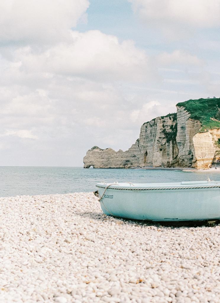 Pebble beach with a pale blue boat in the foreground, facing the calm sea and dramatic white cliffs of Étretat in Normandy, France, under a partly cloudy sky.